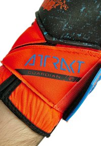 Guanto sportivo arancione con imbottitura nera texturizzata. Presenta il marchio blu "ATRKT GUARDIAN" e una sezione elastica al polso per una vestibilità aderente.