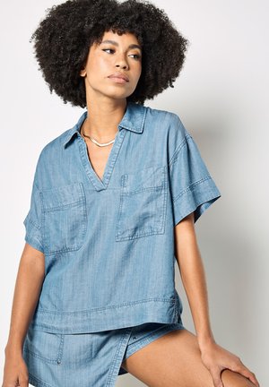 Kobieta o kręconych włosach nosi luźną niebieską koszulę dżinsową z dwoma kieszeniami na piersiach oraz dopasowane spodenki, siedząc na jasnym tle.