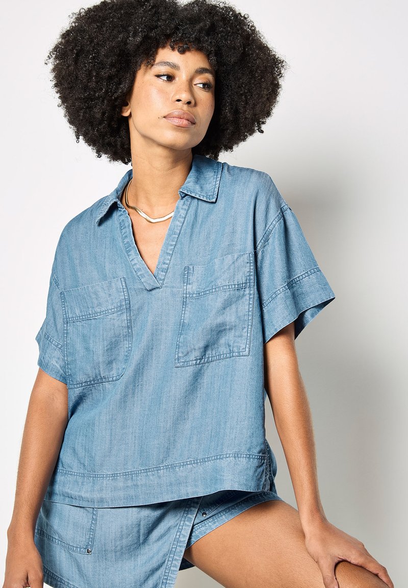Femme aux cheveux bouclés portant une chemise en denim bleu ample avec deux poches poitrine et un short assorti, assise devant un fond clair.