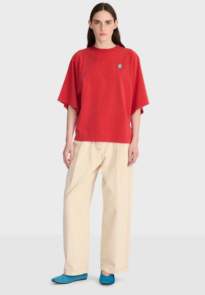 Femme portant un haut rouge ample à manches courtes, un pantalon large beige et des chaussures plates bleues, debout contre un fond clair uni.