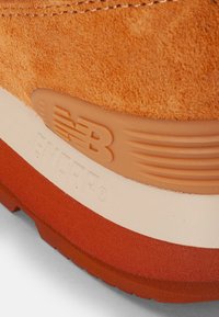 Close-up of orange suede sneaker heel with embossed New Balance logo and layered rubber sole in tan, white, and red tones.