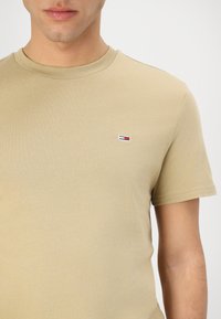 T-shirt in cotone beige con maniche corte e colletto rotondo. Presenta un piccolo logo tricolore sul lato sinistro del petto. Texture liscia.