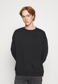 Sweatshirt noir en tissu doux, présentant un col rond et des épaules tombantes. La coupe est décontractée, rehaussée de poignets et d'un ourlet côtelés.