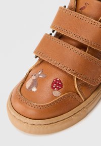 Zapatilla de cuero marrón con dos correas de velcro. Presenta diseños bordados de conejo y hongo. Punta redondeada y suela de goma.