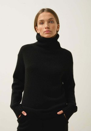 Schwarzer Rollkragenpullover aus weichem Strickmaterial, mit lockerer Passform, gerippten Bündchen und Saum, während die Hände in den Taschen liegen.