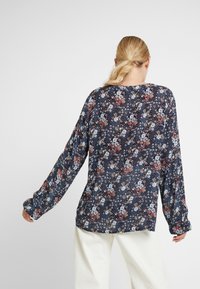 Blumenbluse mit langen Ärmeln in Dunkelblau, die eine Mischung aus roten, blauen und cremefarbenen Blumen zeigt, locker geschnitten und mit weicher Textur.
