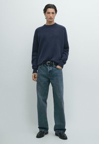 Marineblauer Strickpullover mit Rundhalsausschnitt und gerippten Bündchen, kombiniert mit locker sitzender dunkelblauer Jeans aus Denim und schwarzen Lederschuhen.