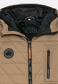 Beige quiltet jakke med sort inderside, udstyret med et logo-patch, to lynlås-lommer og kontrasterende lynlås-detaljer.