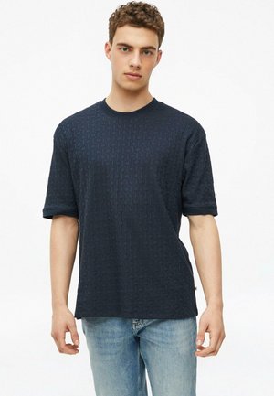 Jeune homme aux cheveux courts et bouclés portant une chemise sombre texturée à manches courtes et un jean bleu clair, debout devant un fond blanc.