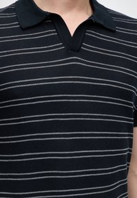 Άνδρας που φοράει μαύρο πόλο με λευκές οριζόντιες ζιγκ-ζαγκ ρίγες και ανοιχτό γιακά, ορατός ο άνω κορμός.