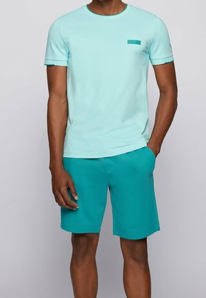 Helles blaues T-Shirt mit kurzen Ärmeln und einer tealfarbenen Akzenttasche, kombiniert mit tealfarbenen Shorts. Beide Teile haben eine glatte Textur und eine lockere Passform.