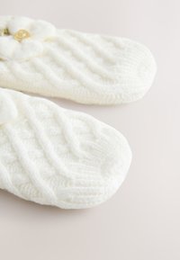 Fehér kötött papucsok mintás textúrával, virágos díszítéssel és arany gomb részlettel a felső részen.