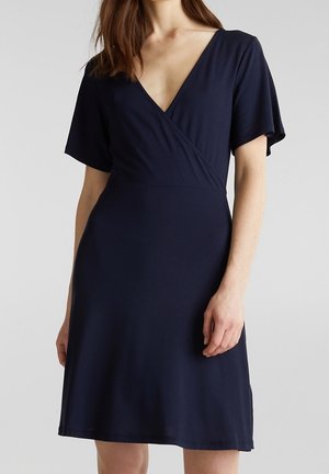 Frau trägt ein marineblaues Kleid mit kurzen Ärmeln, V-Ausschnitt und Wickelstil-Oberteil vor einem schlichten hellen Hintergrund.