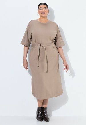 Kvinde med mørkt hår i en beige midi-kjole med bælte og sorte ankelstøvler, stående og smilende mod en hvid baggrund.