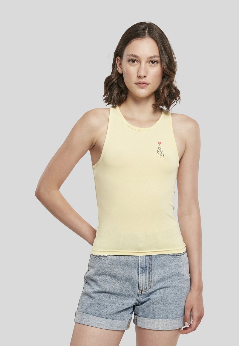 Jeune femme aux cheveux bruns ondulés porte un haut sans manches jaune pâle avec un petit motif main et cœur, associé à un short en jean bleu clair roulé.
