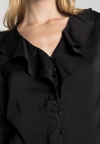 Sort bluse med en flæsekrave, der har en knapdetalje fortil. Fremstillet af glat stof med en blød tekstur.