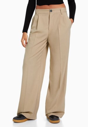 Pantaloni beige a gamba larga con vita alta, pieghe frontali, chiusura con bottone e tasche laterali, abbinati a scarpe nere e beige.