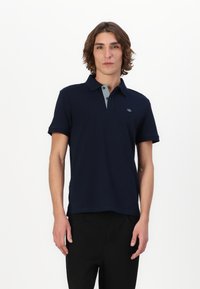 Tricou polo bleumarin din bumbac, cu guler gri deschis și mâneci scurte. Logo mic pe piept. Purtați cu pantaloni negri.