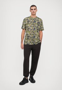 Short-sleeve t-shirt features a multicolor camouflage pattern in green, yellow, and blue. Paired with dark pants and black sneakers.