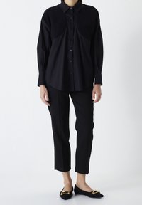 Persona in piedi che indossa una camicia nera con bottoni e pannelli textured sulle spalle, pantaloni neri a vita alta e ballerine nere con dettagli di fibbia dorata.