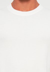 Close-up of a person wearing a plain white ribbed crew neck shirt, showing neck and upper chest area.