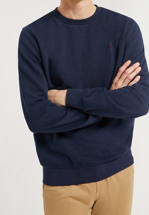 Hombre con los brazos cruzados vestido con una sudadera azul marino de cuello redondo y pantalones marrón claro sobre un fondo liso.