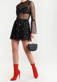 Schwarzes durchsichtiges Oberteil mit Blumenstickerei, schwarzer Rock, rote Stiefeletten und eine kleine schwarze Handtasche mit rundem Griff.