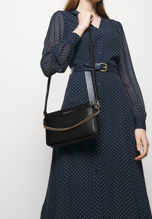 Czarna torba na ramię z łańcuszkowym akcentem, wykonana z gładkiego materiału, zestawiona z granatową sukienką w kropki z guzikami i paskiem.