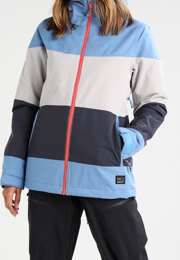 Personne portant une veste bicolore avec des panneaux bleus, gris clair et gris foncé, une fermeture éclair rouge, et un pantalon noir avec des poches zippées.