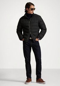 Juoda paminkšta striukė su aukštu apykakliu ir užtrauktuku, dėvima su tamsiomis džinsais ir rudos spalvos batais. Modelis vilki akinius nuo saulės ir stovi pasitikėdamas savimi.