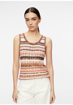 Kobieta o ciemnych włosach ma na sobie bezrękawnik z prążkowanego dzianego materiału z poziomymi paskami w kolorach brązowym, różowym i białym, dopasowany do białych spodni, na jednolitym tle.