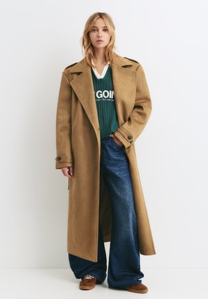 Lange tan kleurige jas met een brede revers, laag over een groene trui en blauwe wijde jeans. Bruine schoenen maken de outfit compleet. Glad textuur.