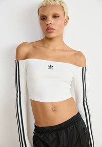 Bijela crop bluzica s jednim ramenom, dugim rukavima crnih pruga i Adidas logotipom na sredini. Tkanina izgleda glatko i usko.