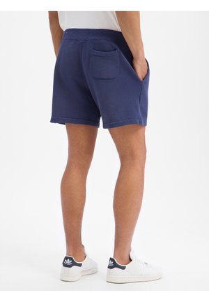 Mann trägt marineblaue Shorts mit Gesäßtasche und weiße Sneaker, steht mit den Händen in den Taschen, mit dem Rücken zur Kamera vor schlichtem Hintergrund.