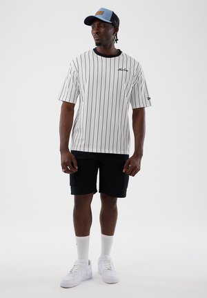 Mann iført en hvit T-skjorte med tynne striper, svarte shorts, hvite sokker, hvite joggesko, og en blå og svart baseballcaps står mot en ensfarget bakgrunn.