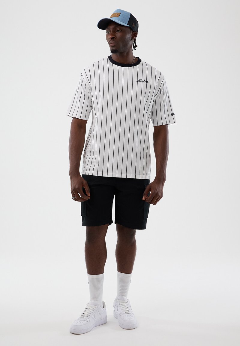 Άνδρας που φοράει λευκό μπλουζάκι με λεπτές ρίγες, μαύρο σορτς, λευκές κάλτσες, λευκά αθλητικά παπούτσια και μπλε-μαύρο καπέλο μπέιζμπολ, στέκεται μπροστά σε απλό φόντο.