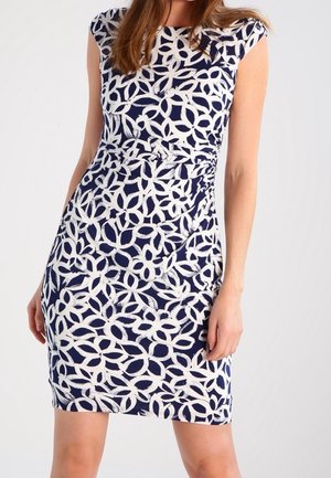 Femme portant une robe ajustée longueur genoux avec un motif abstrait de feuilles blanc et bleu marine, debout devant un fond uni.