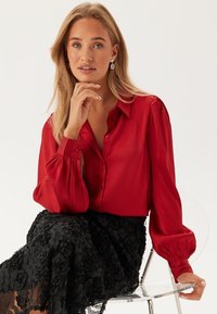 Rode blouse met lange mouwen en een kraag, uitgevoerd in een textuurstof en met een knoopsluiting. Gepaard met een zwarte rok, versierd met bloemdetails.