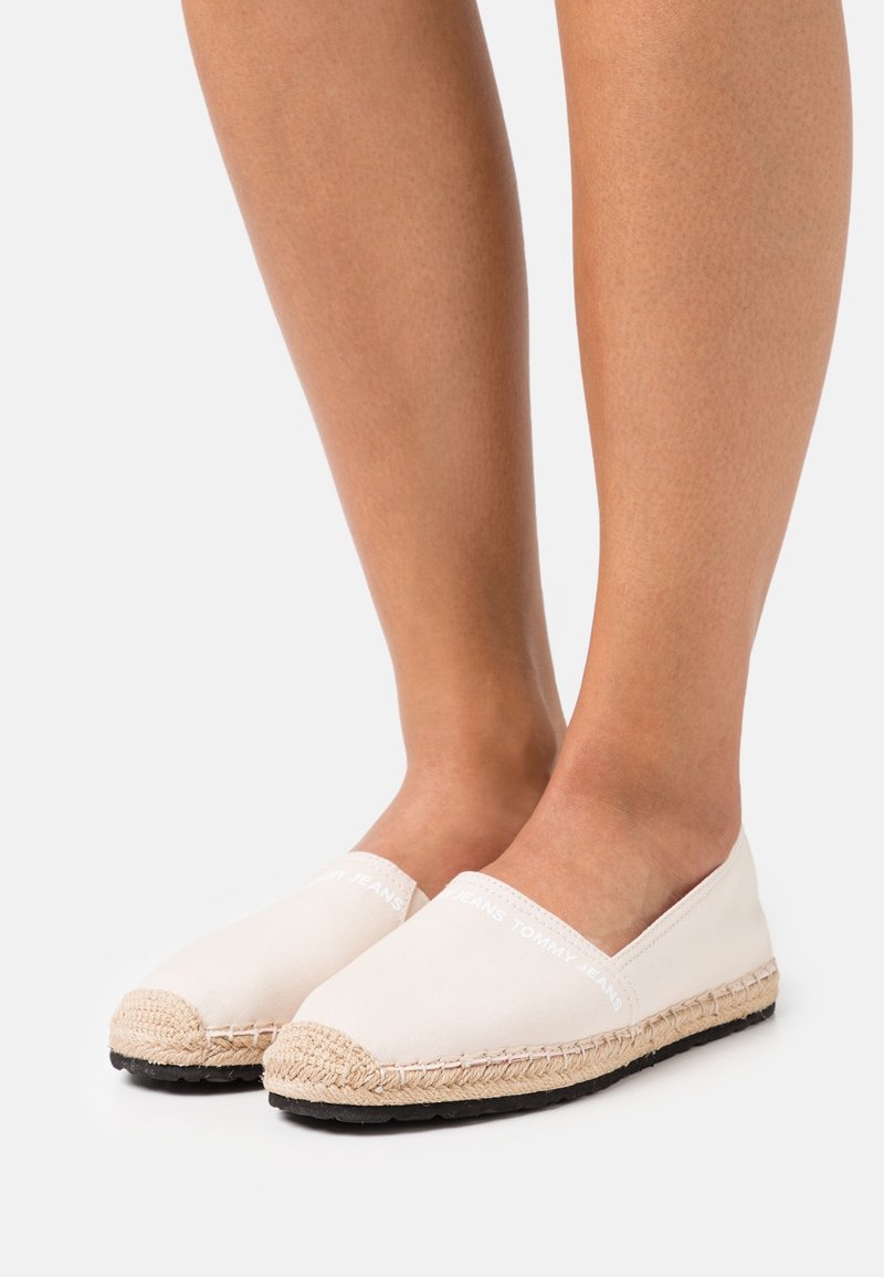 Espadrilles en toile beige clair avec une semelle en jute tissée et un fond en caoutchouc noir. Présente un logo de marque blanc sur le devant. Design classique à enfiler.