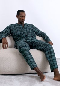 Πράσινο και μαύρο καρό σετ λούτρου με μπλούζα χωρίς γιακά και παντελόνι ίσιας γραμμής. Μαλακό ύφασμα και χαλαρή εφαρμογή, φοριέται ενώ κάθεται σε μια μπεζ καρέκλα.