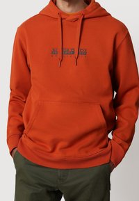 Oranžová mikina s kapucí a přední kapsou typu klokan. Má tónovaný logotyp v modré barvě. Vyrobena z měkké látky s žebrovanými manžetami a lemem.