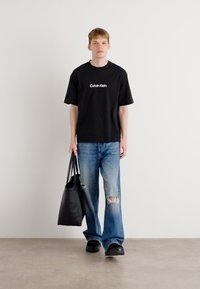 Svart t-skjorte med hvit "Calvin Klein" logo, lyseblå ripte jeans, svart tote bag og svarte sko med tykk såle.