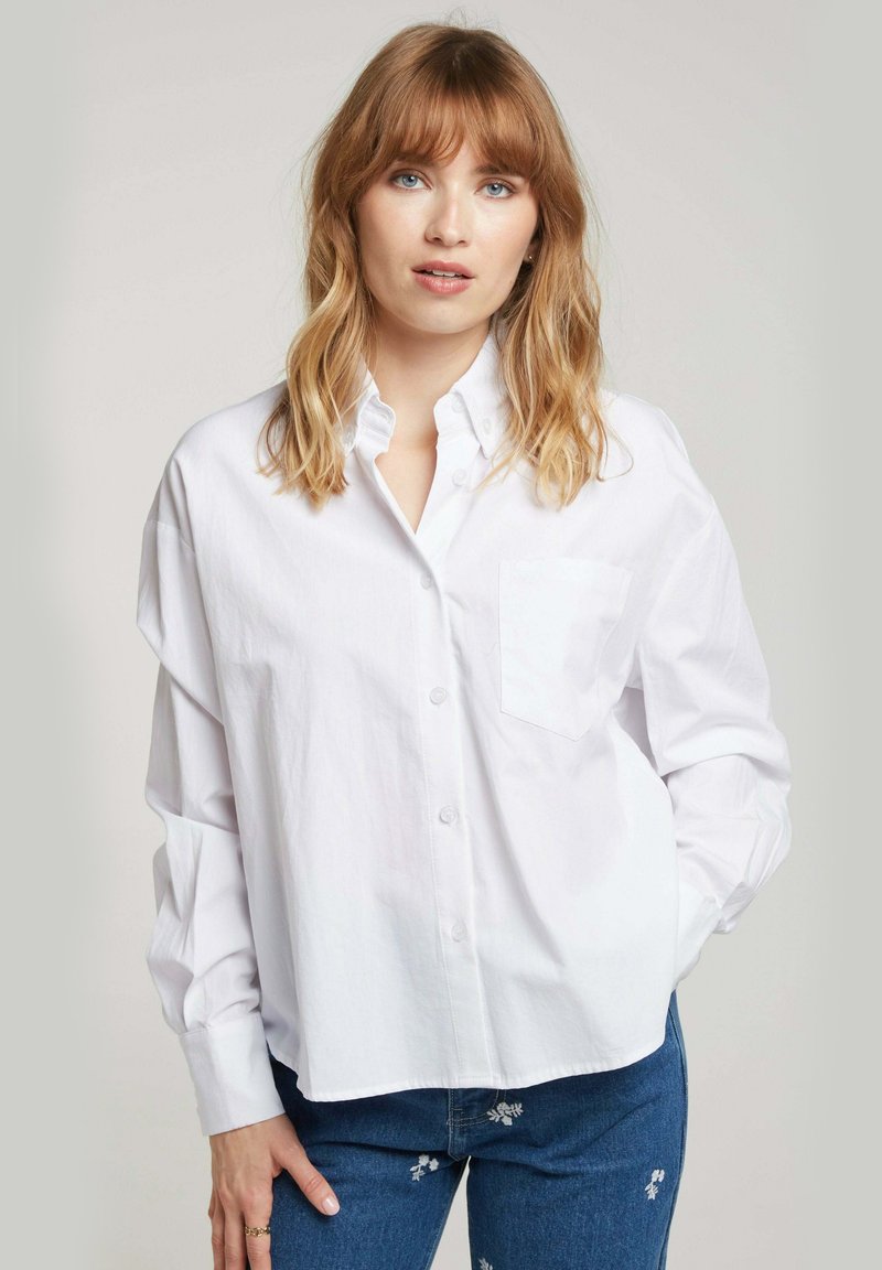 Chemise blanche à boutons avec une poche poitrine simple, manches longues, coupe décontractée et texture lisse. Portée avec un jean bleu brodé.