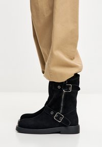 Crne gležnjače od brušene kože s bočnim patentnim zatvaračem, remenom i kopčom, ravnim potplatom, u kombinaciji s kaki hlačama. Tekstura izgleda mekano i mat.