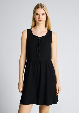 Blonde Frau trägt ein ärmelloses schwarzes Kleid mit rundem Ausschnitt und Bindeband-Details, steht vor einem einfarbigen hellgrauen Hintergrund.