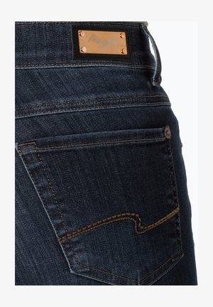 Tummat farkut, joissa on neliömäinen timantti kohokuvioidulla brändilogolla, kuparin väriset niitit ja kultainen ommel takataskun suunnittelussa.