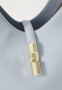 Sac à main en cuir bleu clair avec un fermoir en métal doré brillant portant un logo stylisé. Texture lisse et design à sommet incurvé.