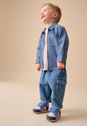Ragazzo giovane che sorride, indossa una giacca blu, camicia bianca, pantaloni di jeans larghi e scarpe da ginnastica colorate, in piedi davanti a uno sfondo beige.