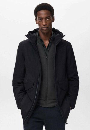 Chaqueta exterior negra con capucha, que presenta un cierre frontal, dos bolsillos laterales y una textura suave. Lleva una capa interna oscura con cremallera debajo.