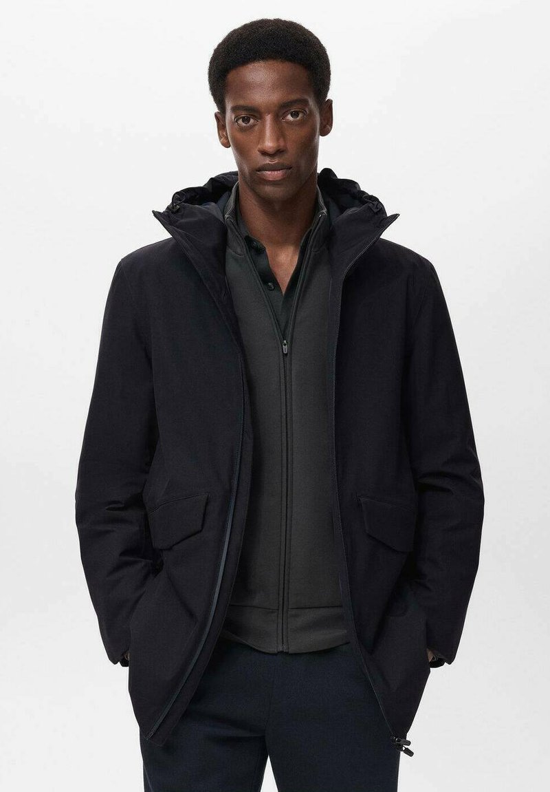 Chaqueta exterior negra con capucha, que presenta un cierre frontal, dos bolsillos laterales y una textura suave. Lleva una capa interna oscura con cremallera debajo.